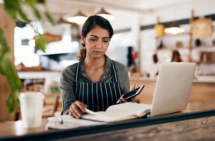 Kvinde gennemgår regnskab i café, laptop, telefon. Ejer af lille virksomhed.