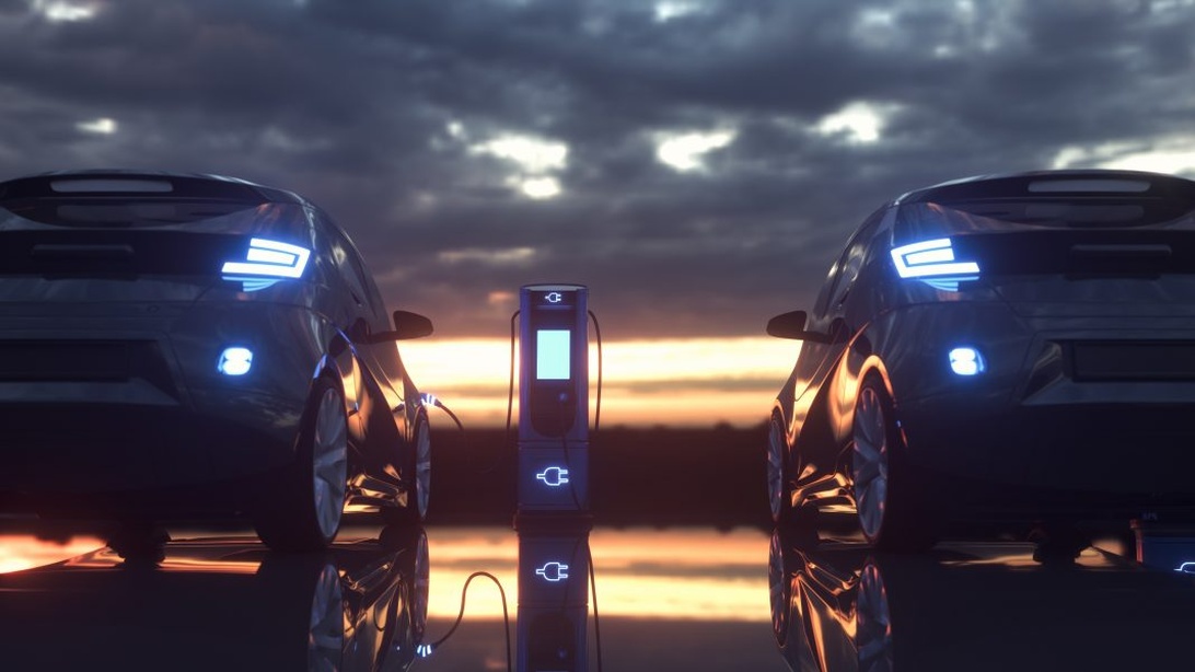 Two electric vehicles parked at a charging station during sunset or sunrise. The dramatic sky with clouds creates a moody atmosphere. The vehicles' headlights are on, and a charging station is visible between them. This appears to be a promotional or artistic shot of EV charging infrastructure.