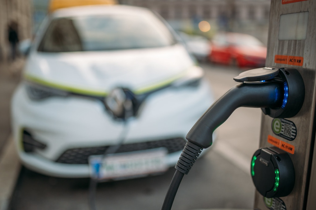 A white Renault electric vehicle connected to a public charging station in an urban setting. The charging cable is prominently visible, and the charging station appears to have indicator lights. The image is taken with a shallow depth of field, keeping the focus on the charging connection while blurring the background.